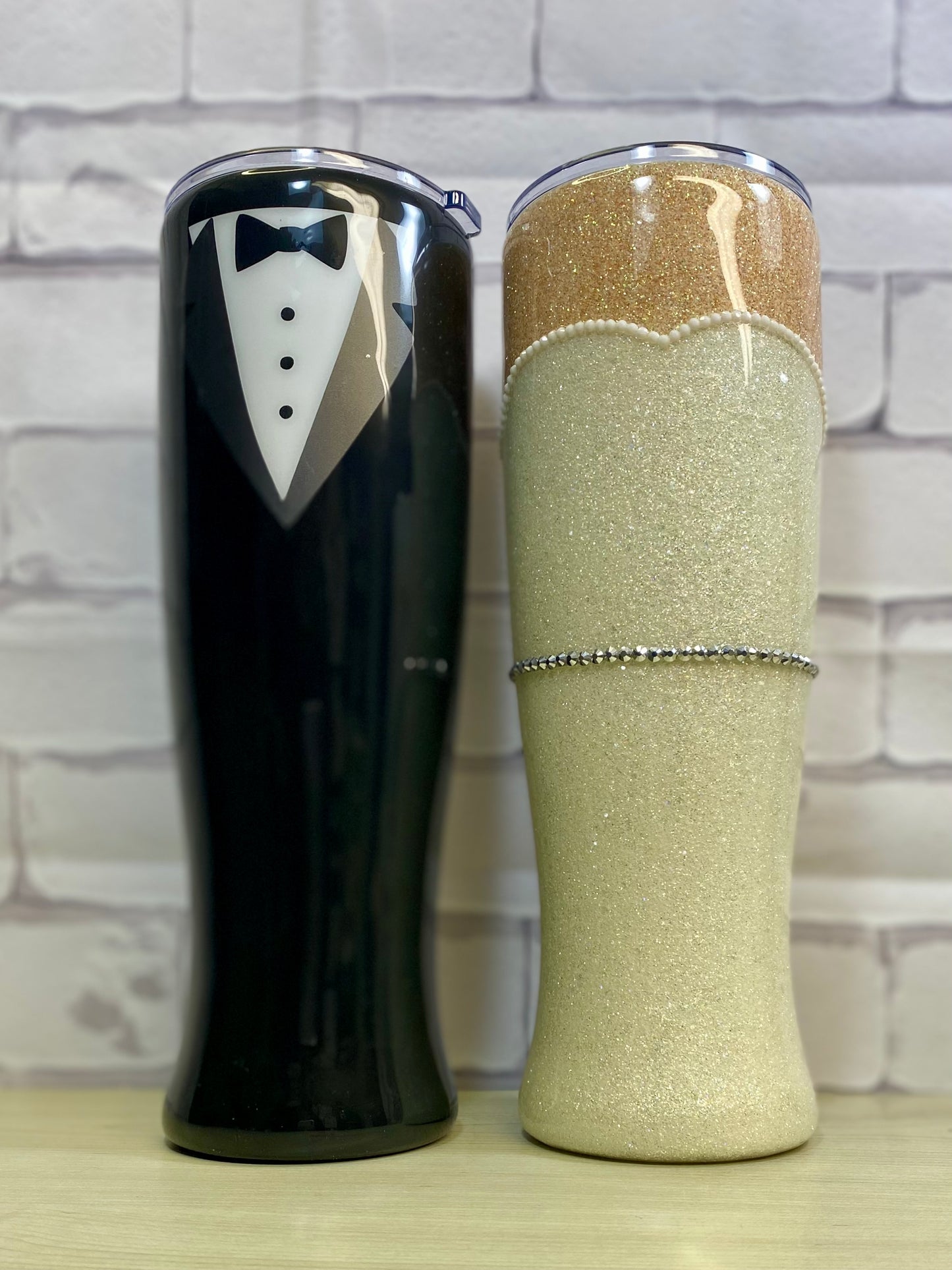 Two stainless steel pilsner tumblers, designed to look like a bride and groom, one black with a white bow tie and lapel detail, and one beige with a brown rim and lace detail.