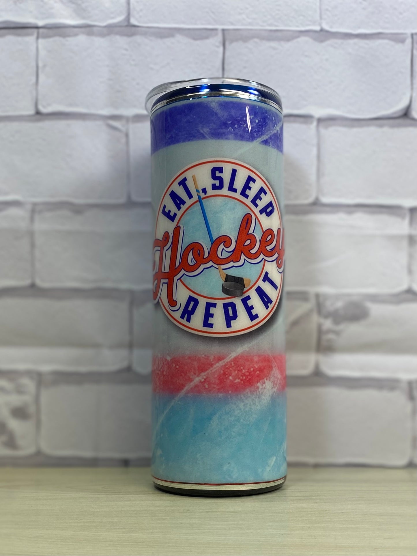 A hockey themed tumbler that looks like an ice rink with the phrase eat, sleep, hockey, repeat on a white brick background.