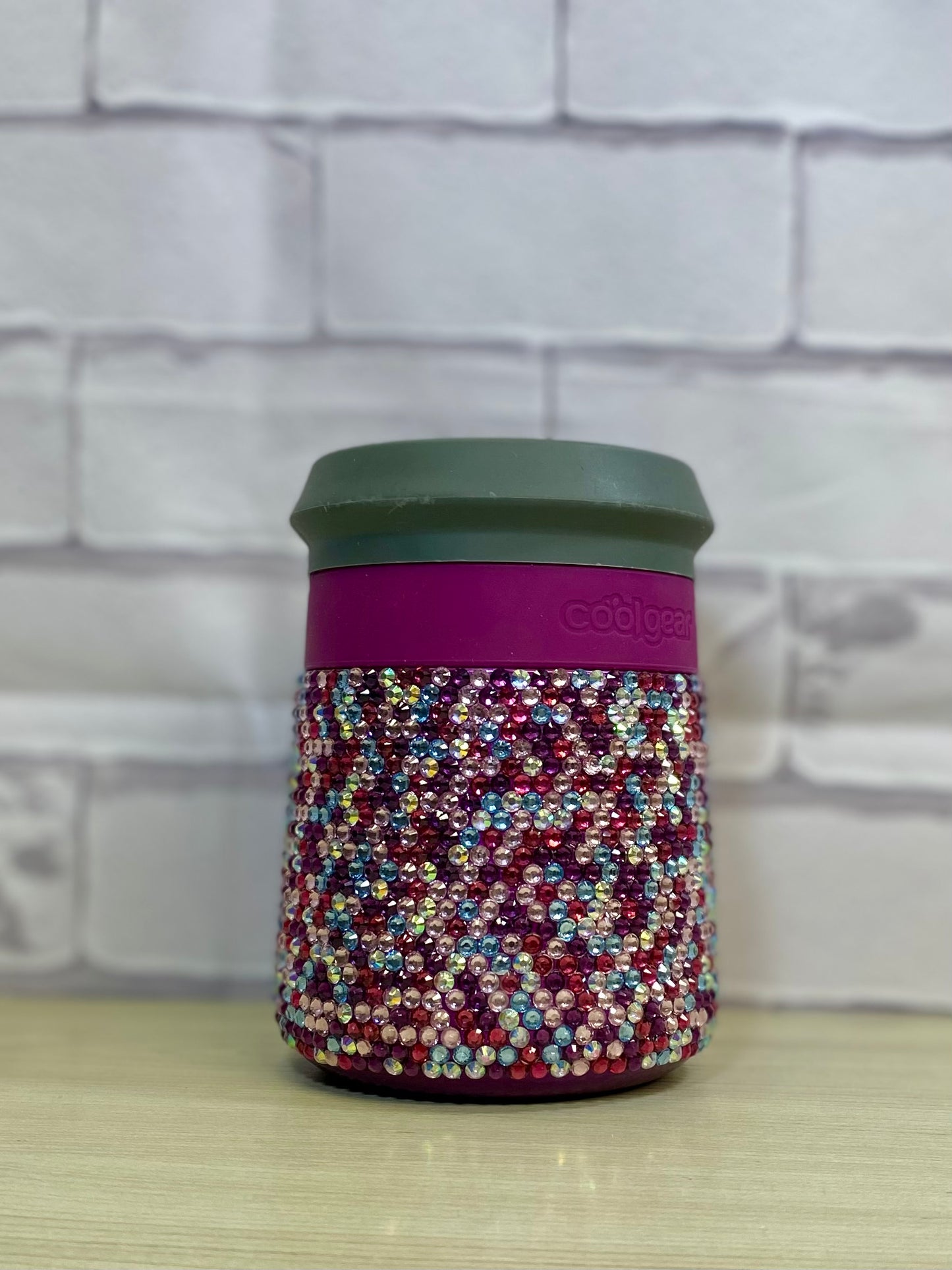 A stainless steel can holder tumbler with a pink rhinestone pattern on a purple background, placed on a wooden surface against a brick wall background.
