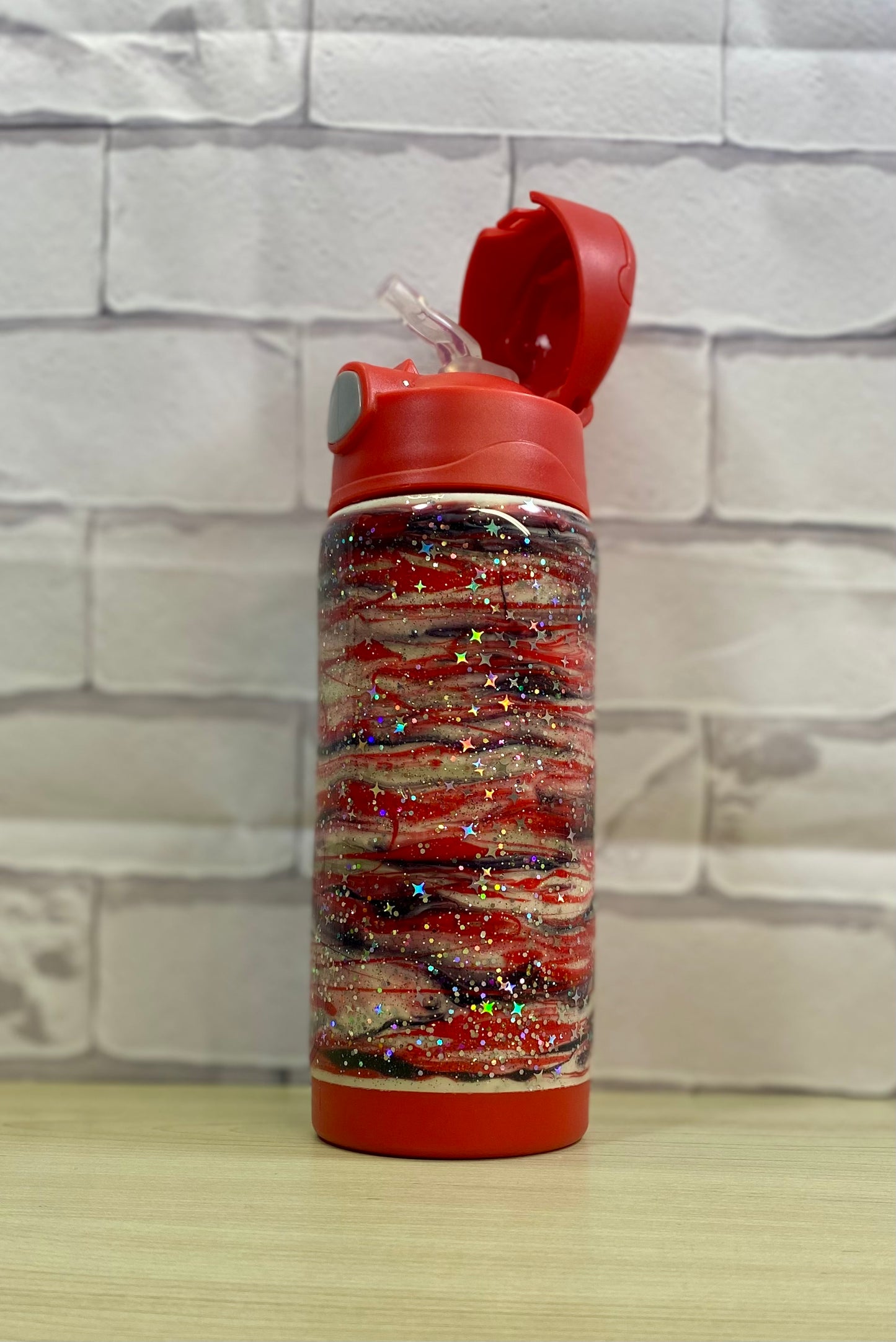 A red glitter swirled kids water bottle with the red lid open, showing a straw with a paisley design on a white brick wall sitting on a wood surface.