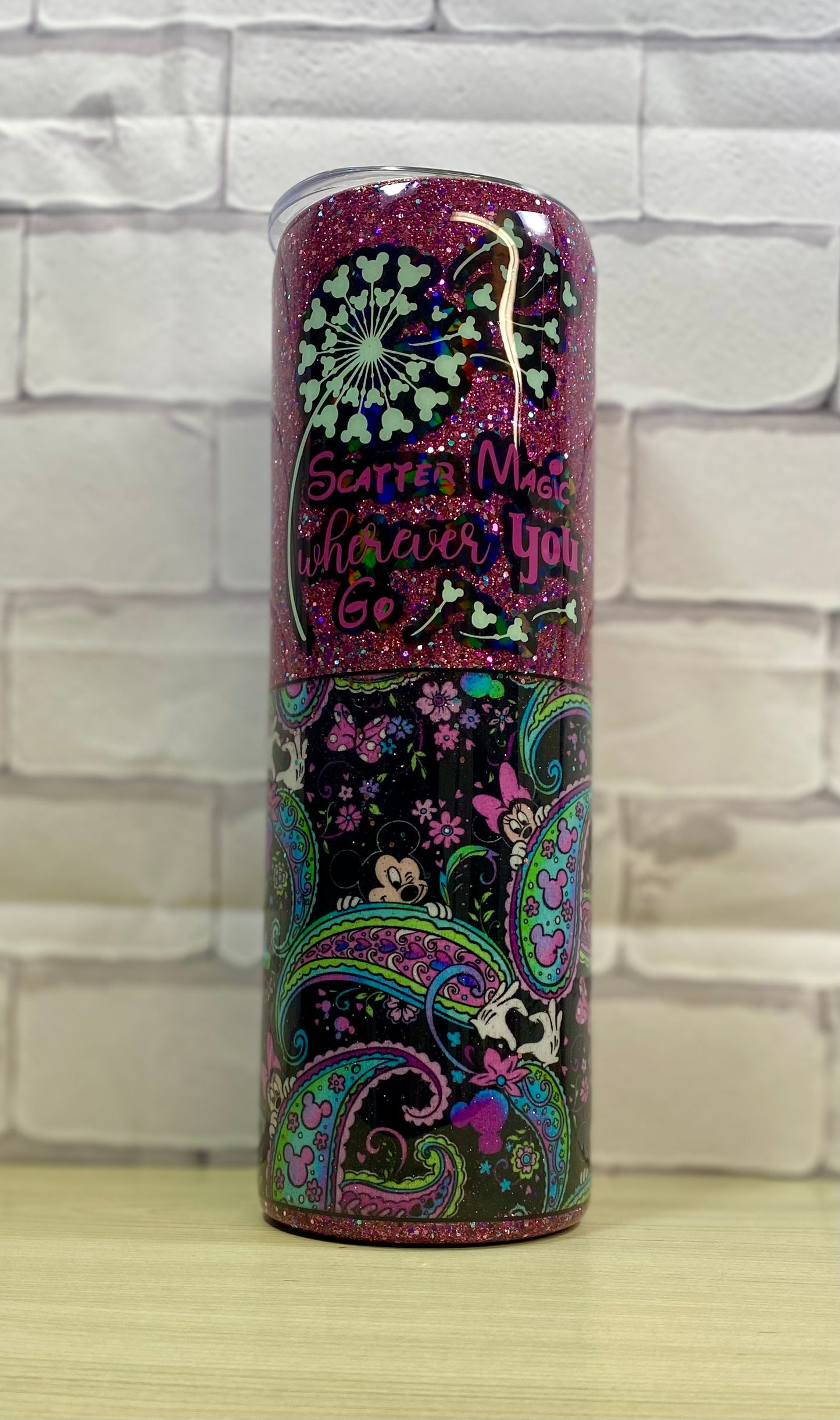 A dark glittered tumbler with a paisley design on a white brick wall sitting on a wood surface.