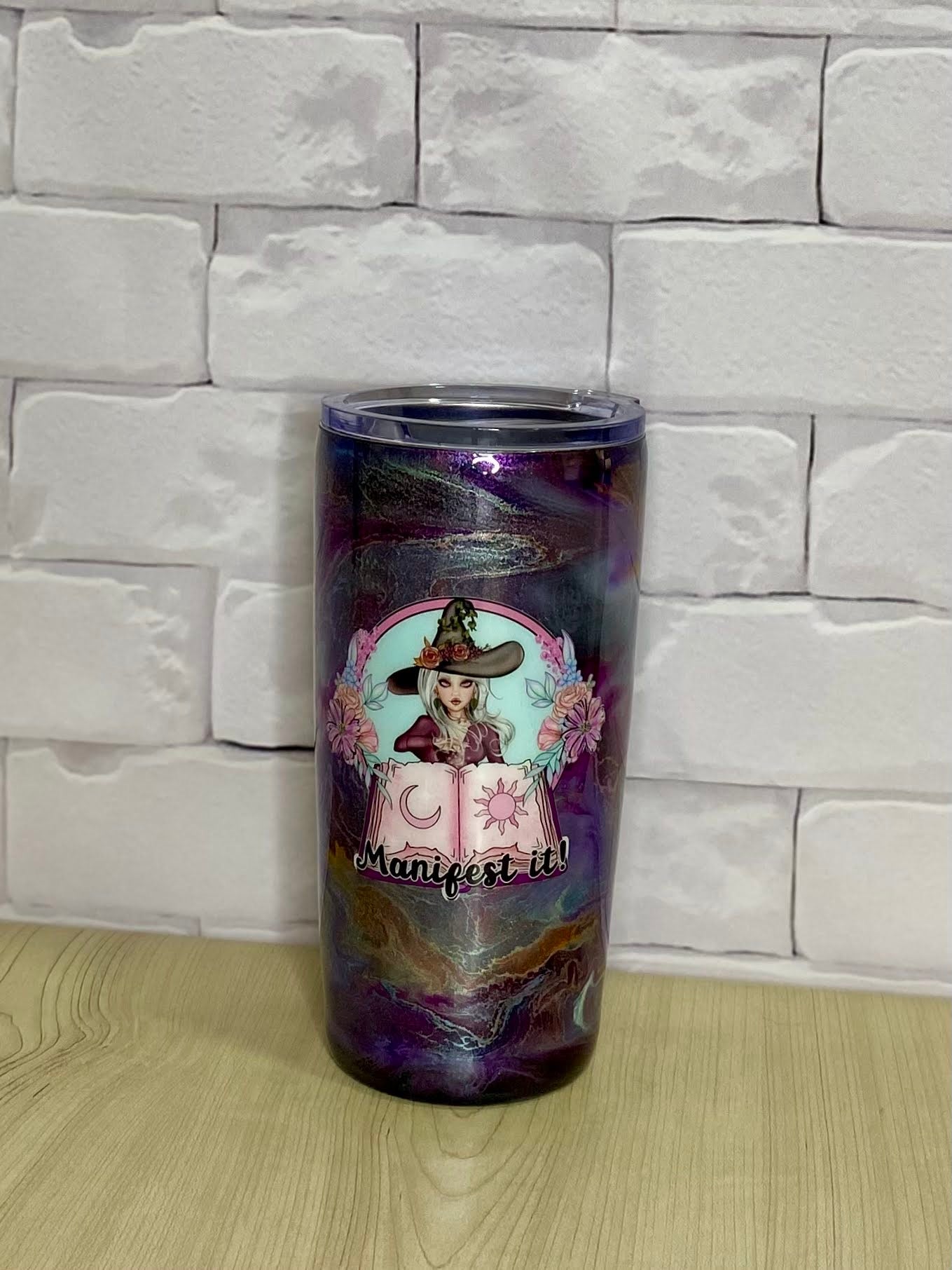 A stainless steel tumbler with a purple and multicolor design, featuring a witch and flowers illustration, placed on a white brick surface.