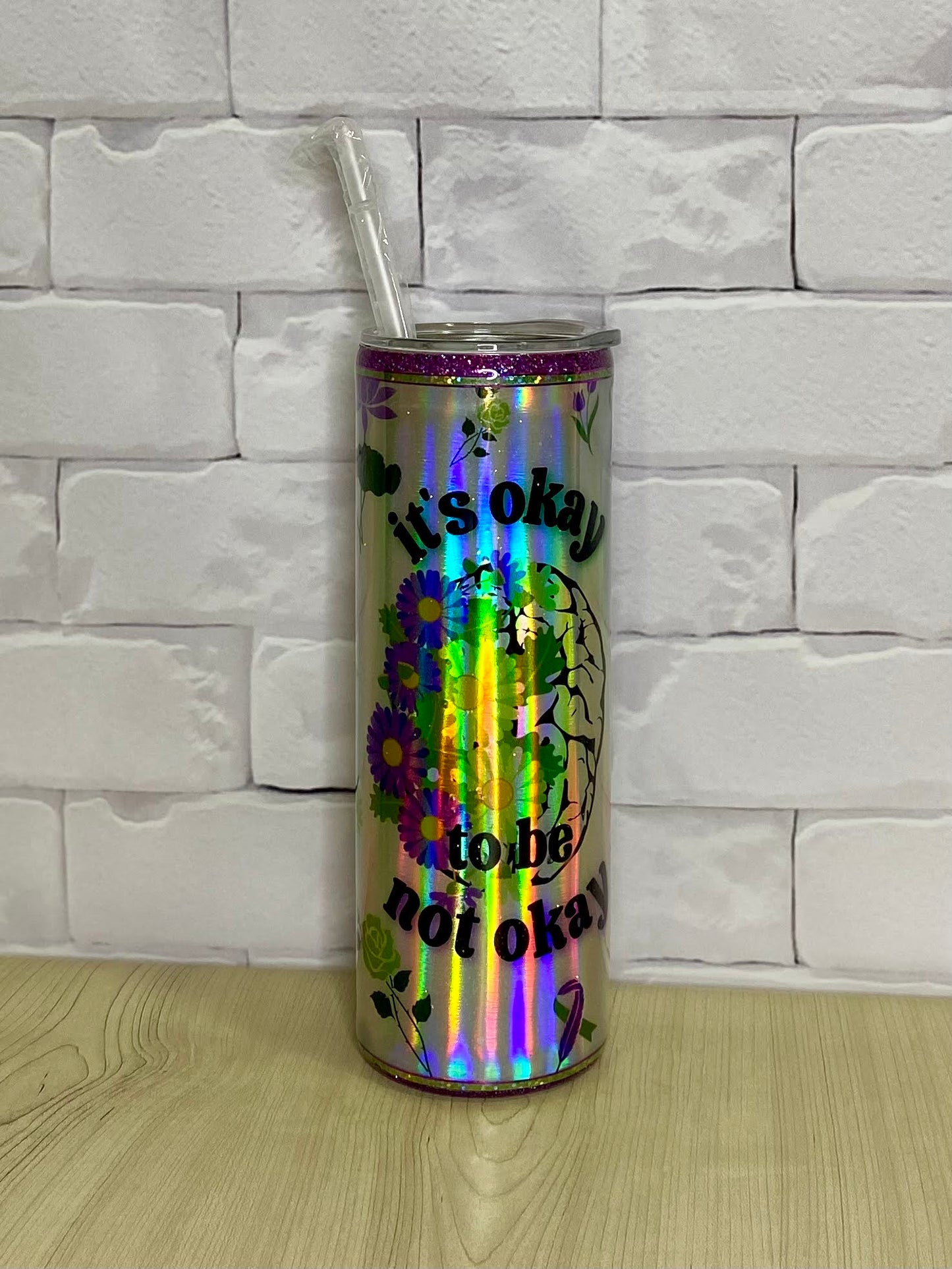 A stainless steel tumbler with a holographic design and the text 'It's OK to not be ok' printed on it, placed on a white surface with a brick wall background.