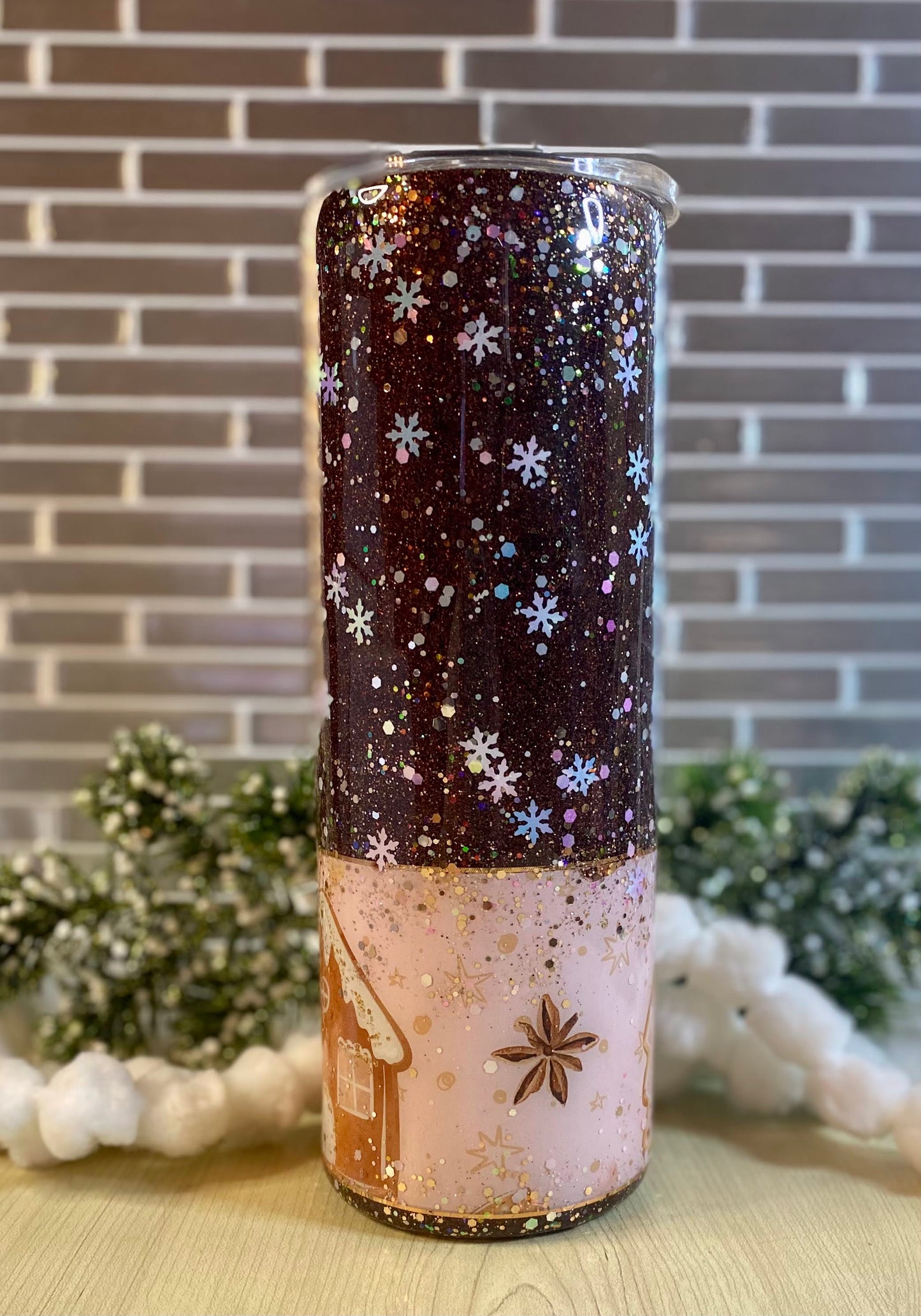A glittered tumbler with snowflakes on a dark glitter and a white glitter bottom with a gold cottage. Displayed on a brick backdrop and sitting on a wooden surface.