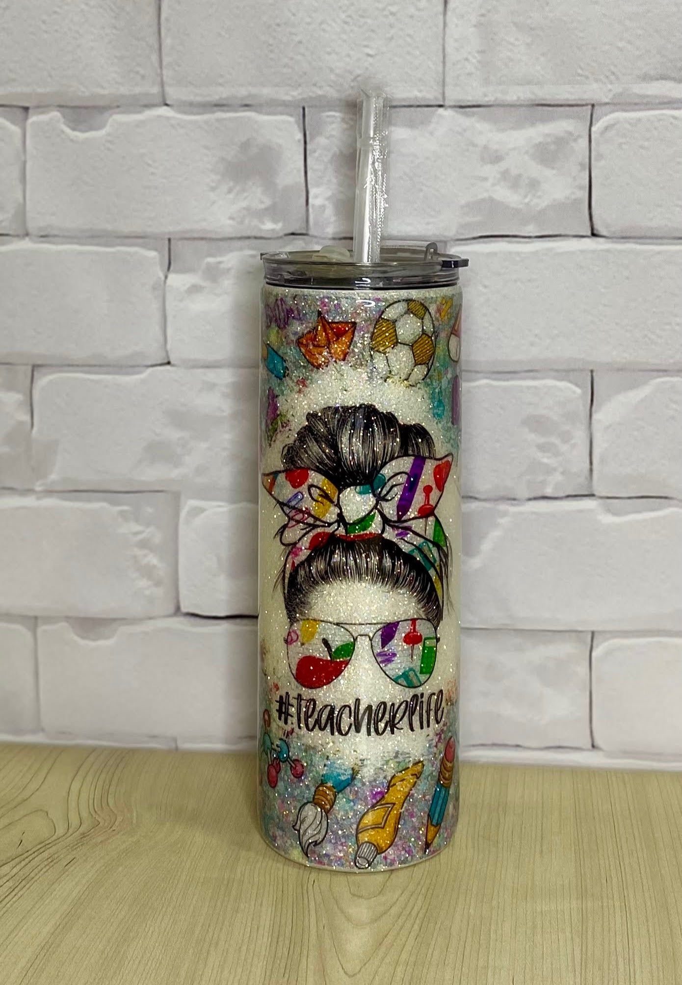 A stainless steel tumbler with a decorative multicolor design, featuring text 'Teacher Life' and various illustrations such as fruits, flowers, and a bow.