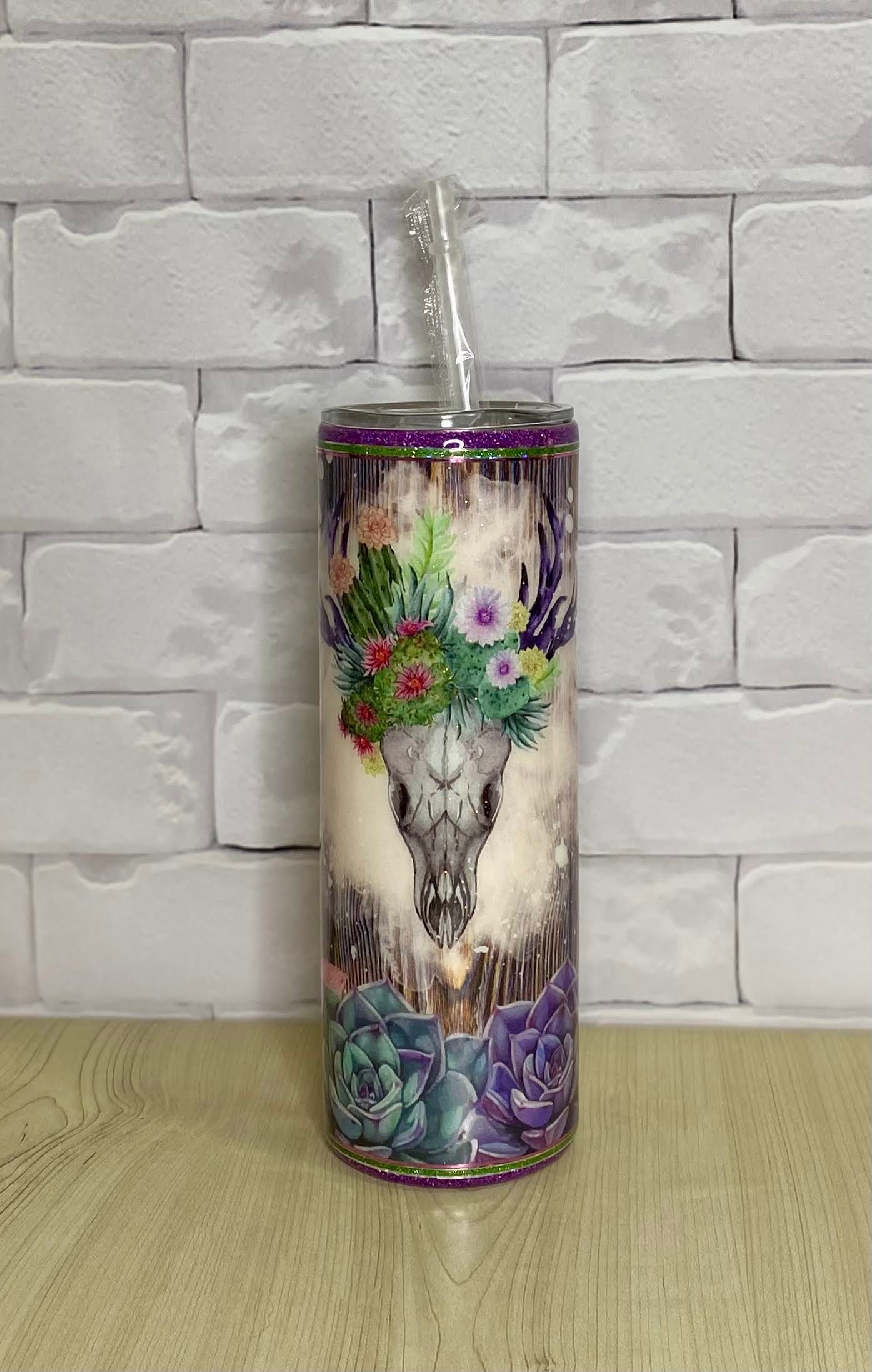 A stainless steel tumbler with a floral and skull design, placed on a white surface with a brick wall background.