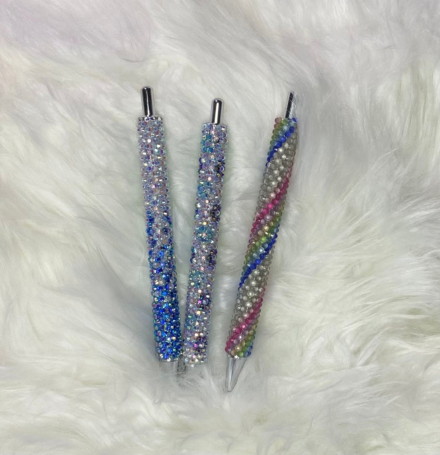 Three custom order rhinestone pens with multicolored rhinestones on the barrel, lying on a white furry surface.
