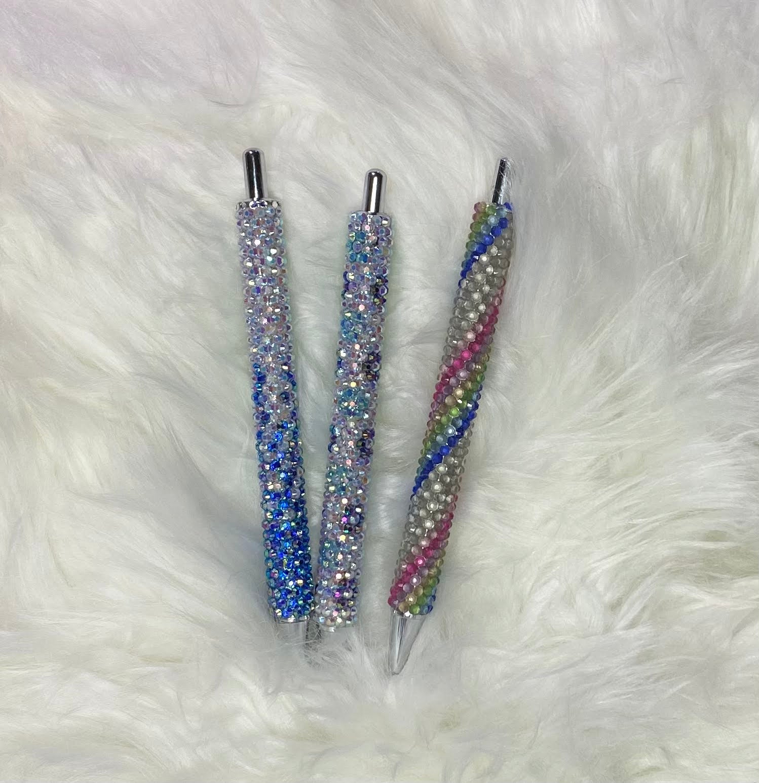 Three custom order rhinestone pens with multicolored rhinestones on the barrel, lying on a white furry surface.